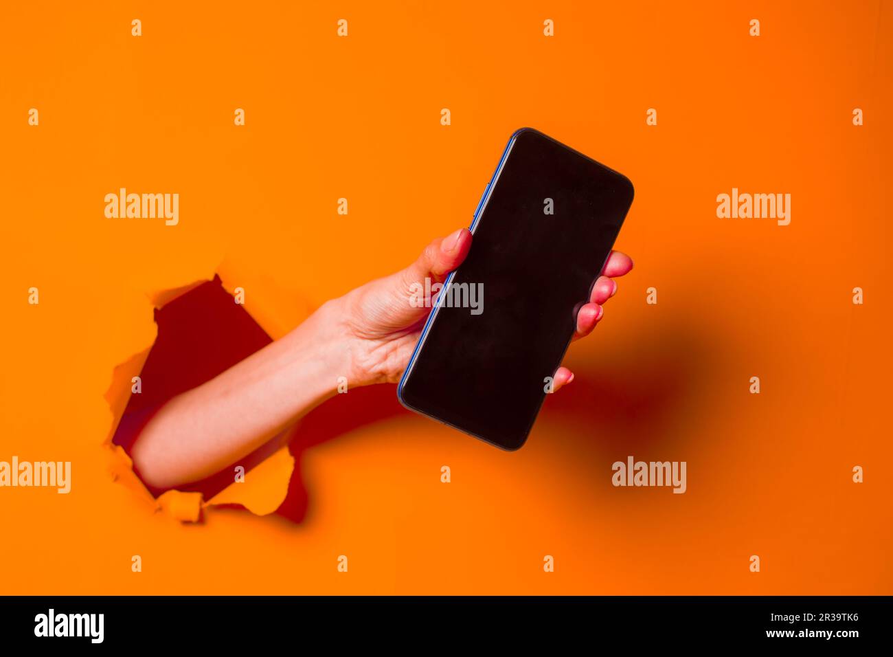 Eine Frau hält ein Telefon durch den verfranste orangefarbene Papierhintergrund, Nahaufnahme. Stockfoto