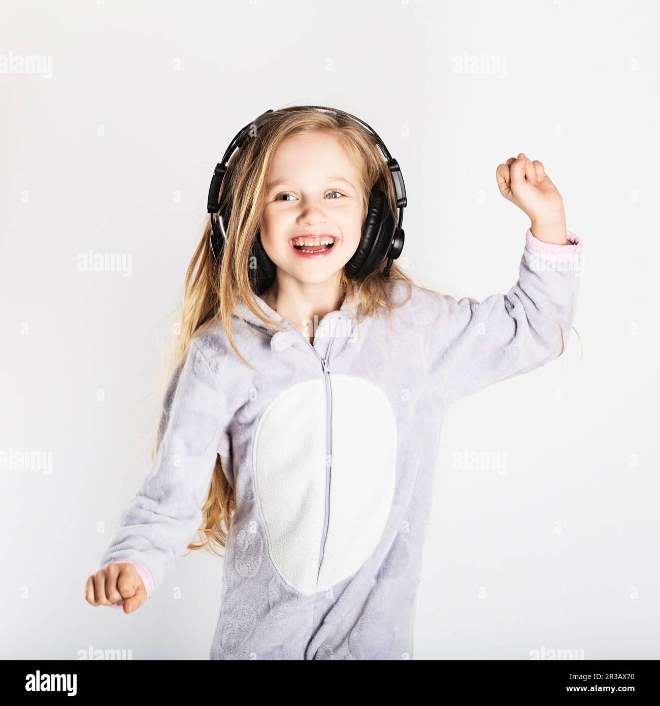 Das süße kleine Mädchen mit Kopfhörern benutzt ein Smartphone, schaut in die Kamera und hat zu Hause Spaß. Tag Stockfoto