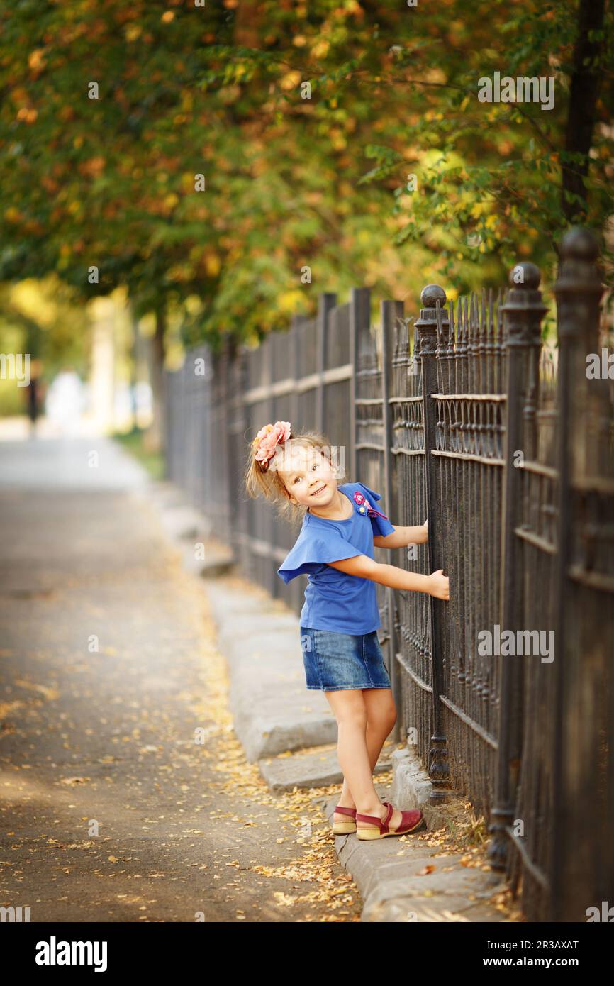 Ein langes Porträt eines kleinen Mädchens, das sich am eisernen Zaun festhält, in einem Stadtpark im warmen Herbst Stockfoto