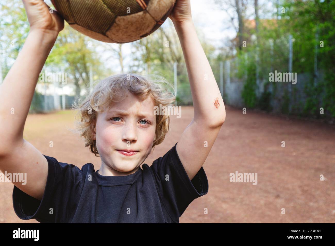 Blonder Junge mit Bluterguss am Unterarm, der Fußball auf dem Spielplatz hält Stockfoto