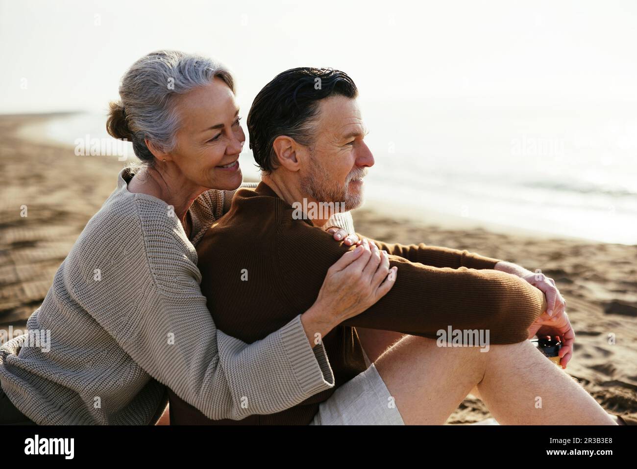 Eine lächelnde Frau umarmt einen Mann, der am Strand sitzt Stockfoto