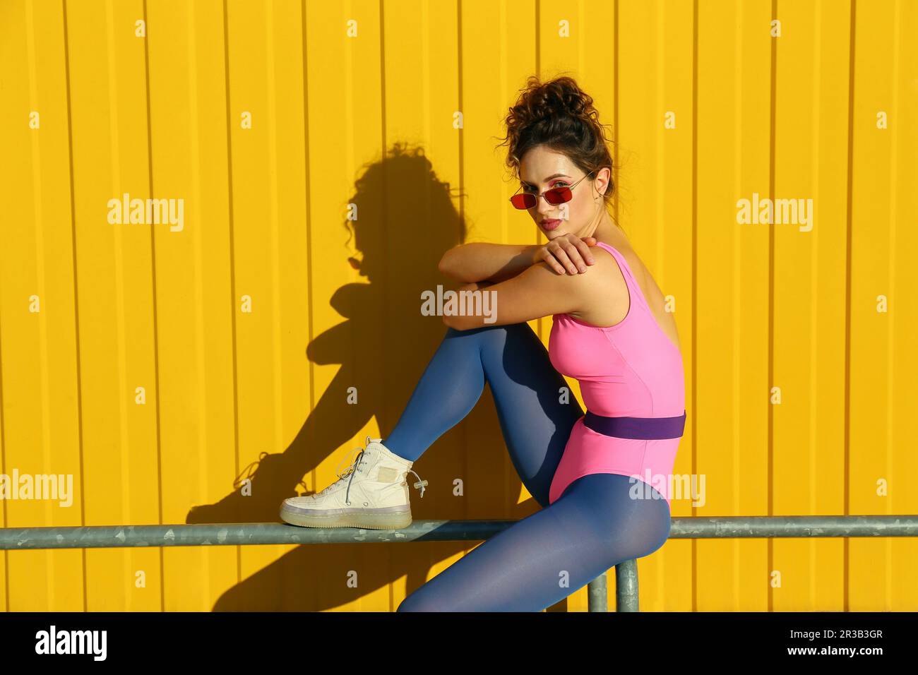 Eine Frau mit Sonnenbrille, die auf dem Geländer vor der gelben Wand sitzt Stockfoto