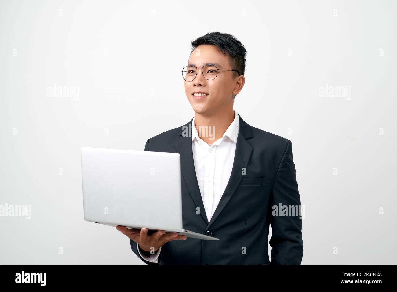 Selbstbewusster junger gutaussehender Mann im schwarzen Anzug, der den grauen Laptop offen hält und beim Stehen lächelt Stockfoto