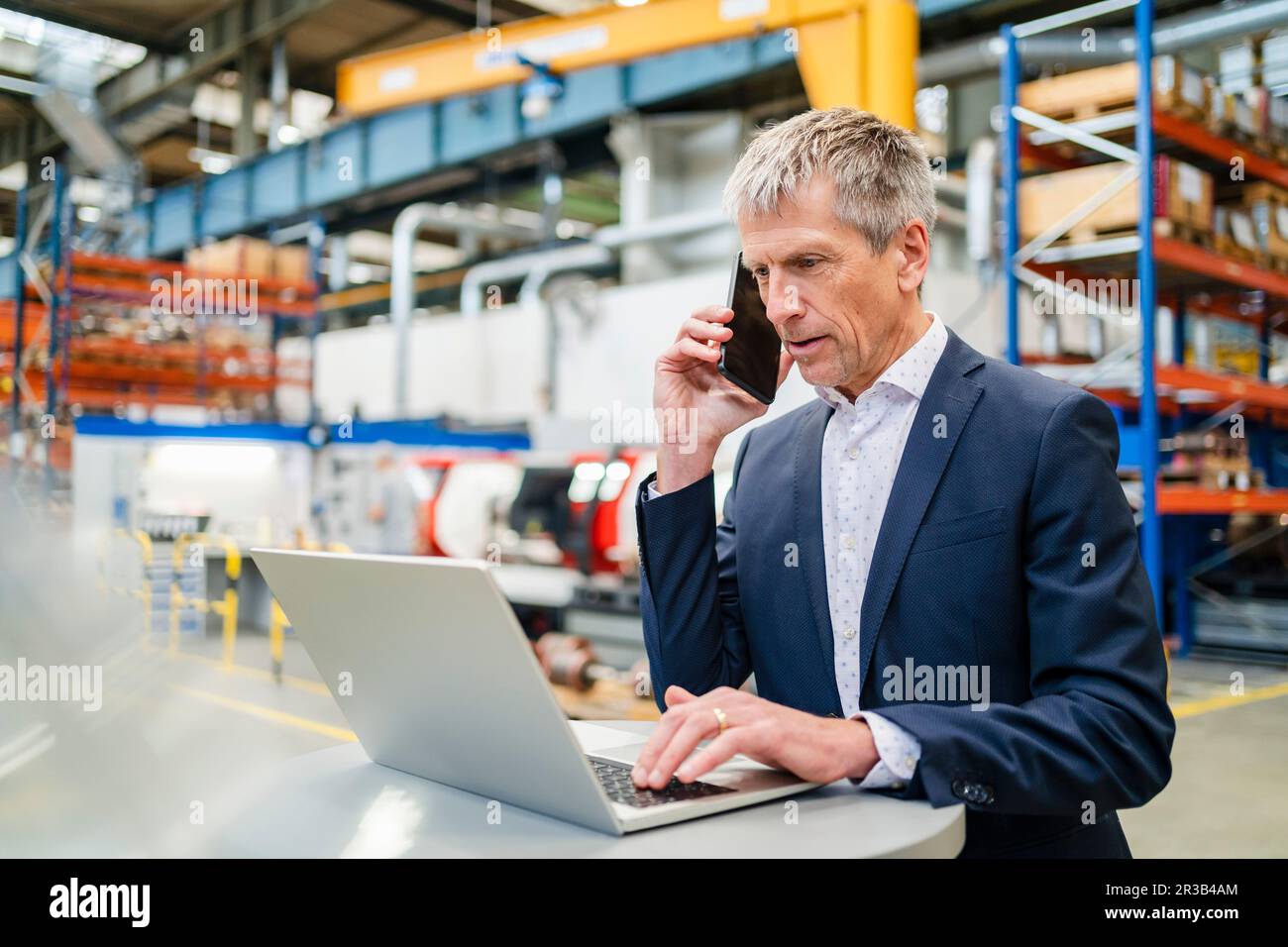 Geschäftsmann, der in der Fabrik mit einem Laptop über ein Smartphone spricht Stockfoto