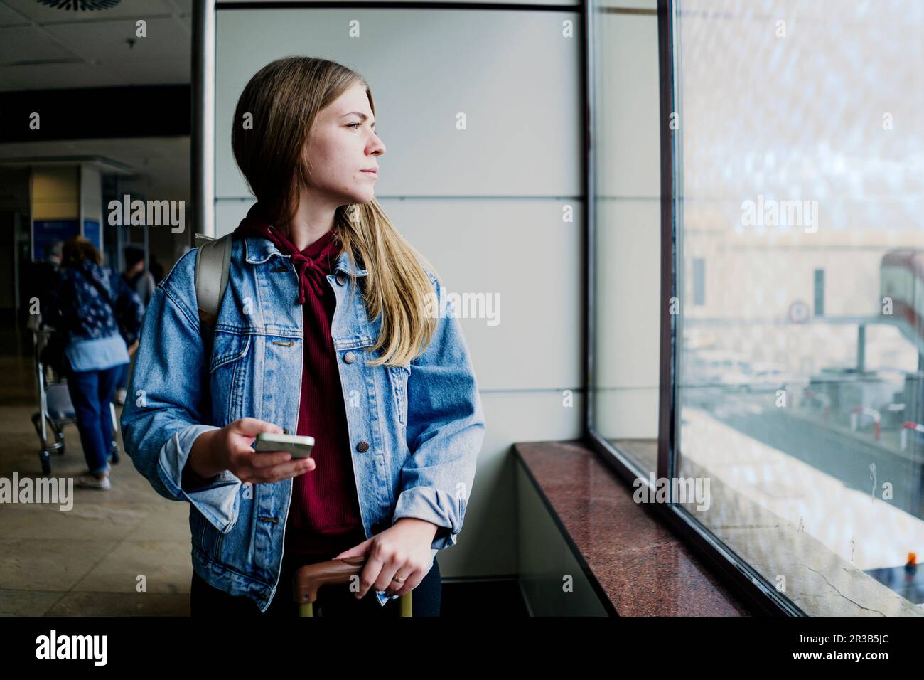 Junge Frau mit Smartphone, die aus dem Fenster am Flughafen schaut Stockfoto