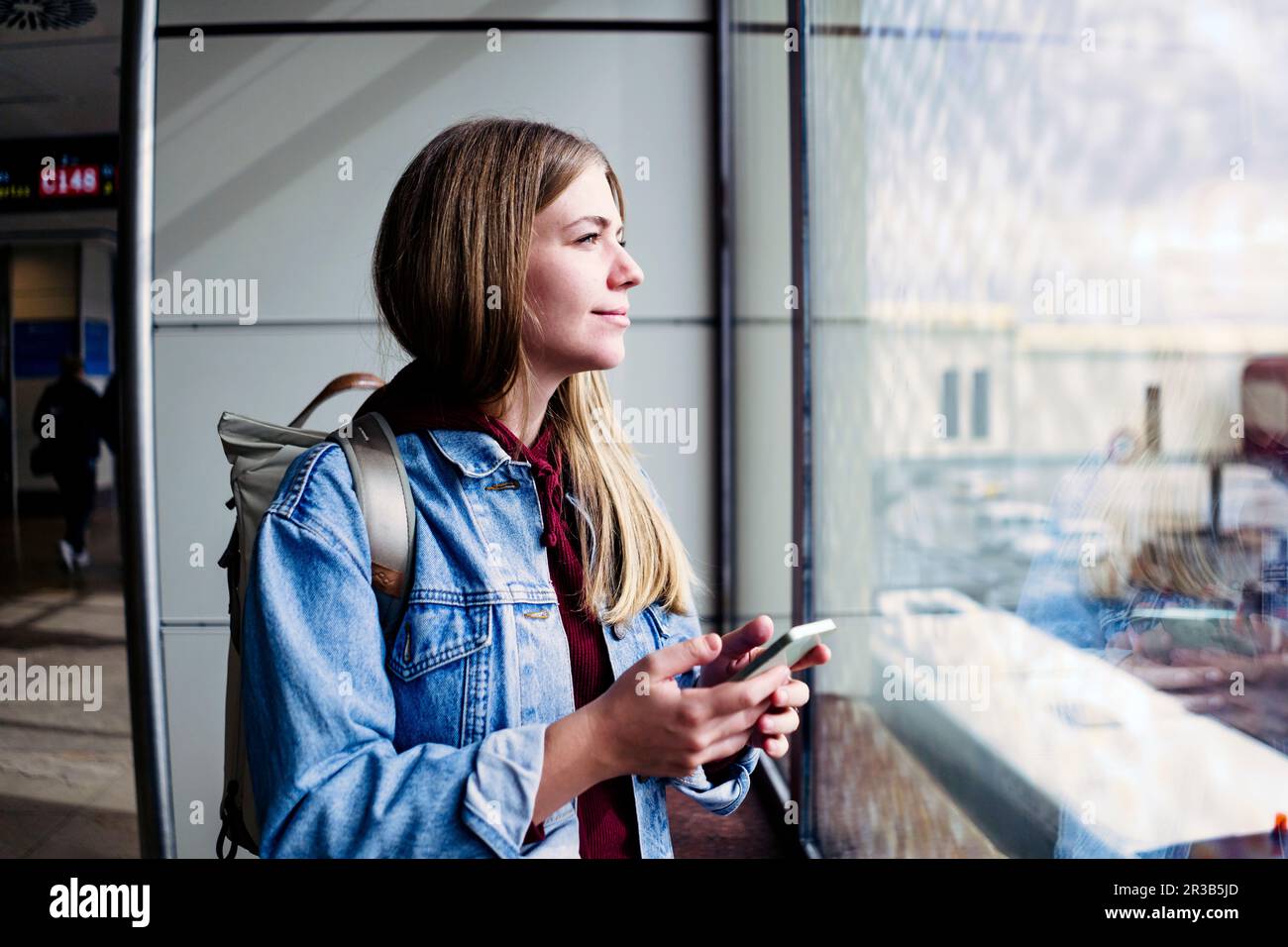 Junge Frau, die aus dem Fenster schaut und ein Smartphone am Flughafen hält Stockfoto