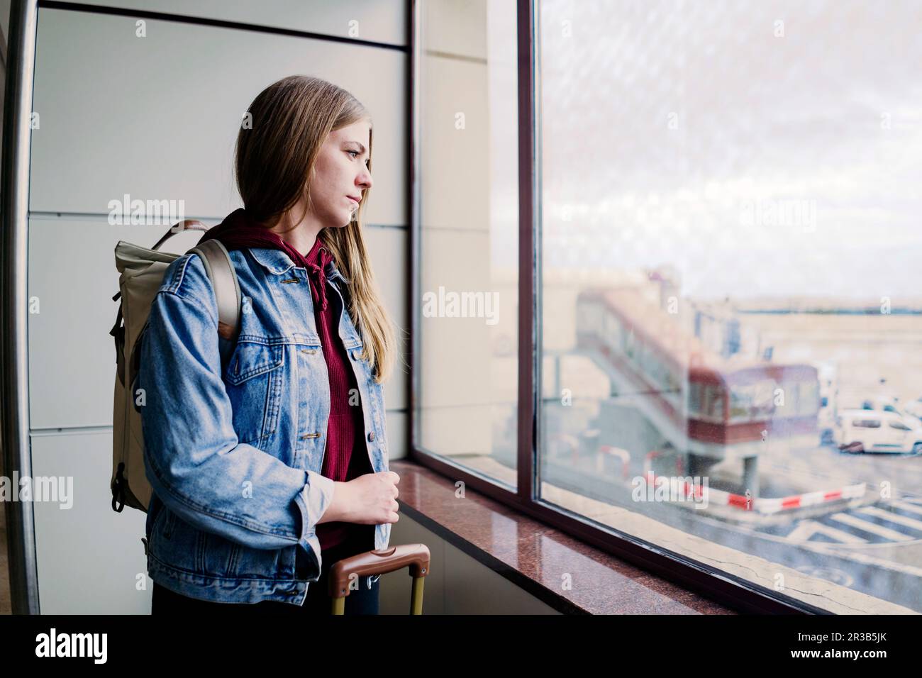 Junge Frau mit Rucksack, die aus dem Flughafenfenster schaut Stockfoto
