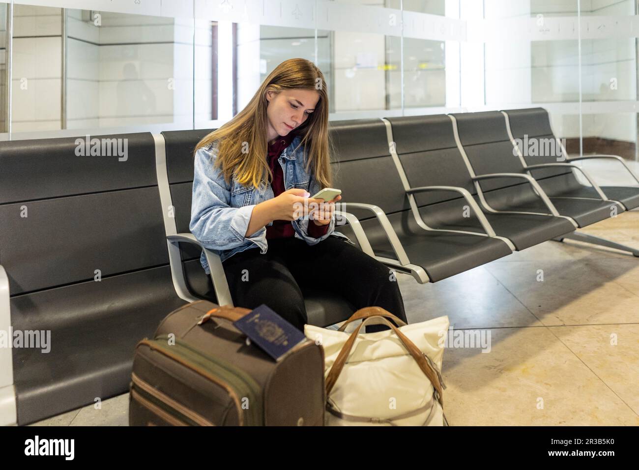 Junge Frau mit Smartphone, die auf dem Sitz in der Flughafenlobby sitzt Stockfoto