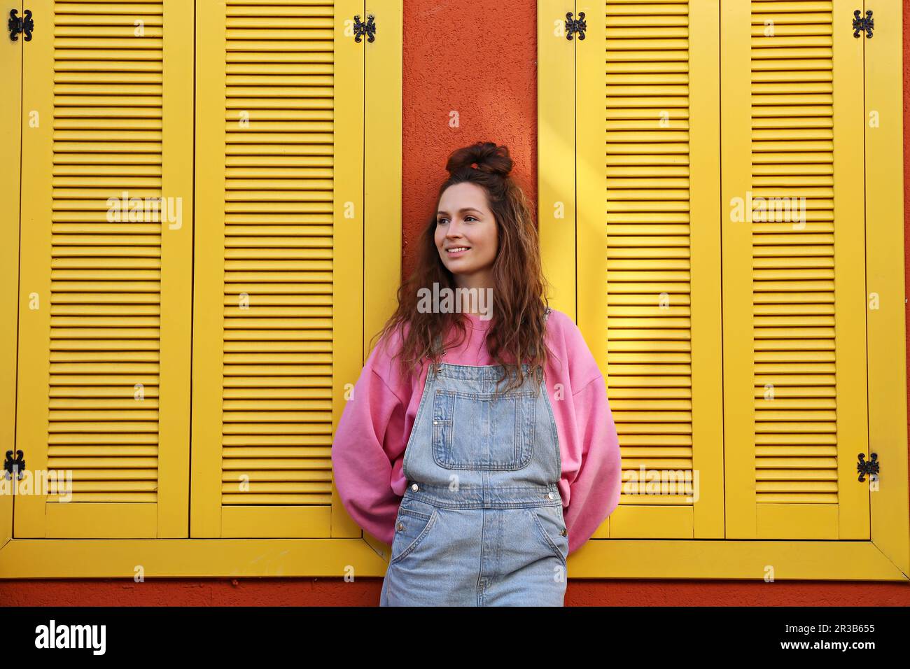 Lächelnde Frau mit Overall, die an der Wand steht, mit gelben Fenstern Stockfoto