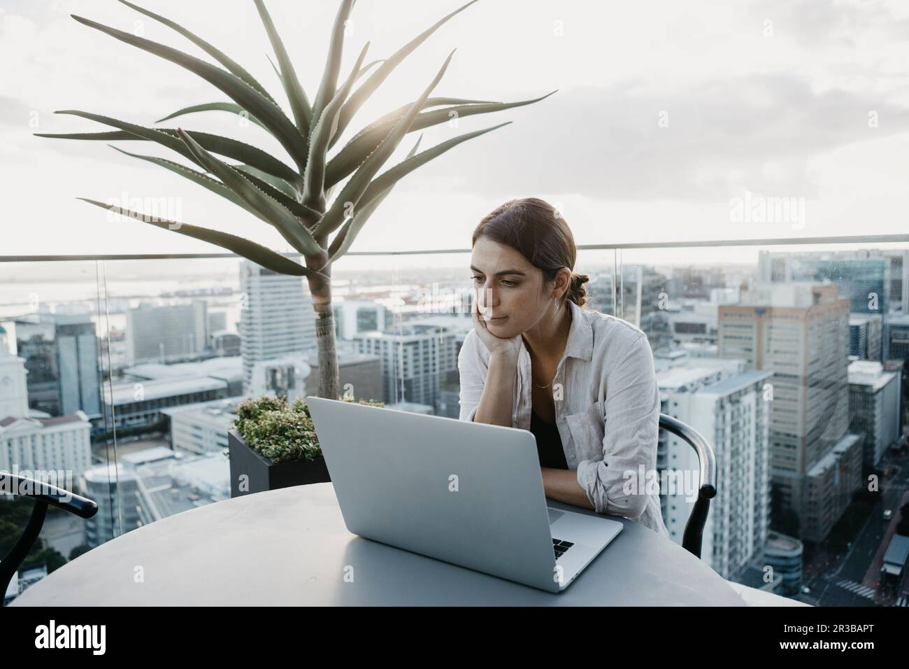 Eine Frau mit der Hand am Kinn, die mit einem Laptop auf dem Dach sitzt Stockfoto