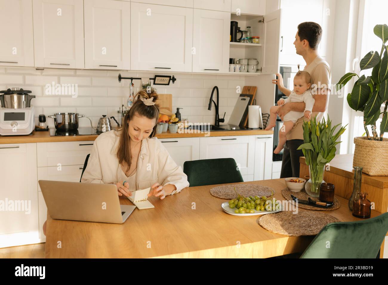 Frau, die an einem Laptop arbeitet, mit Mann, der eine Tochter trägt und zu Hause im Hintergrund arbeitet Stockfoto