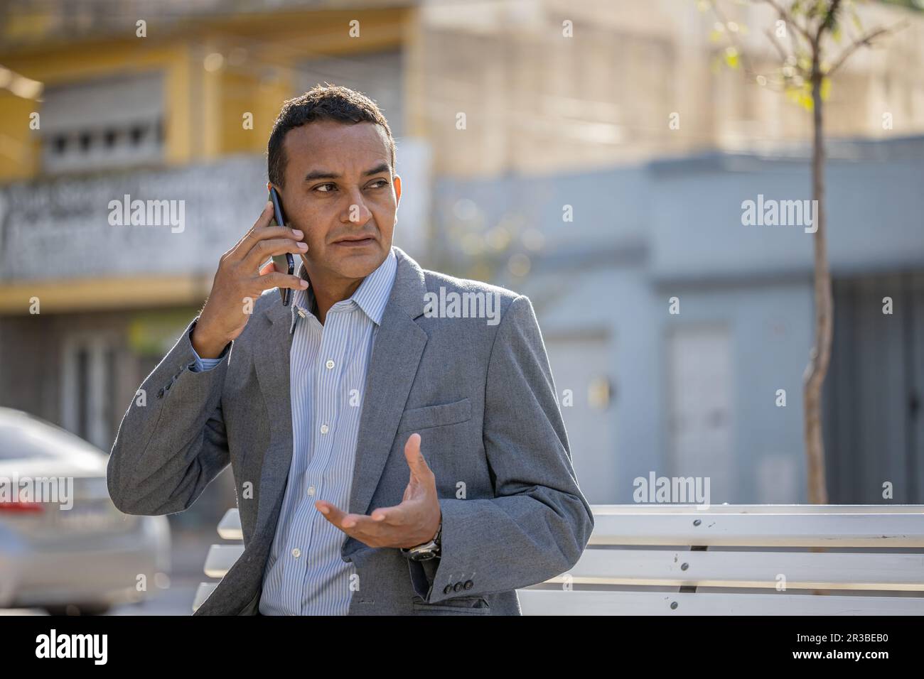 Ein junger latino, der auf einer quadratischen Bank sitzt und mit dem Handy spricht. Stockfoto