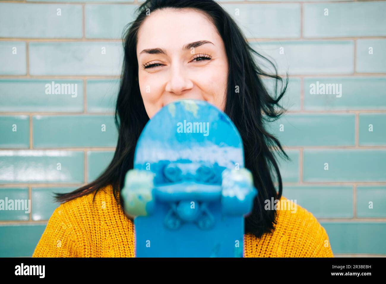 Glückliche junge Frau mit schwarzem Haar, die blaues Skateboard vor der Wand hält Stockfoto