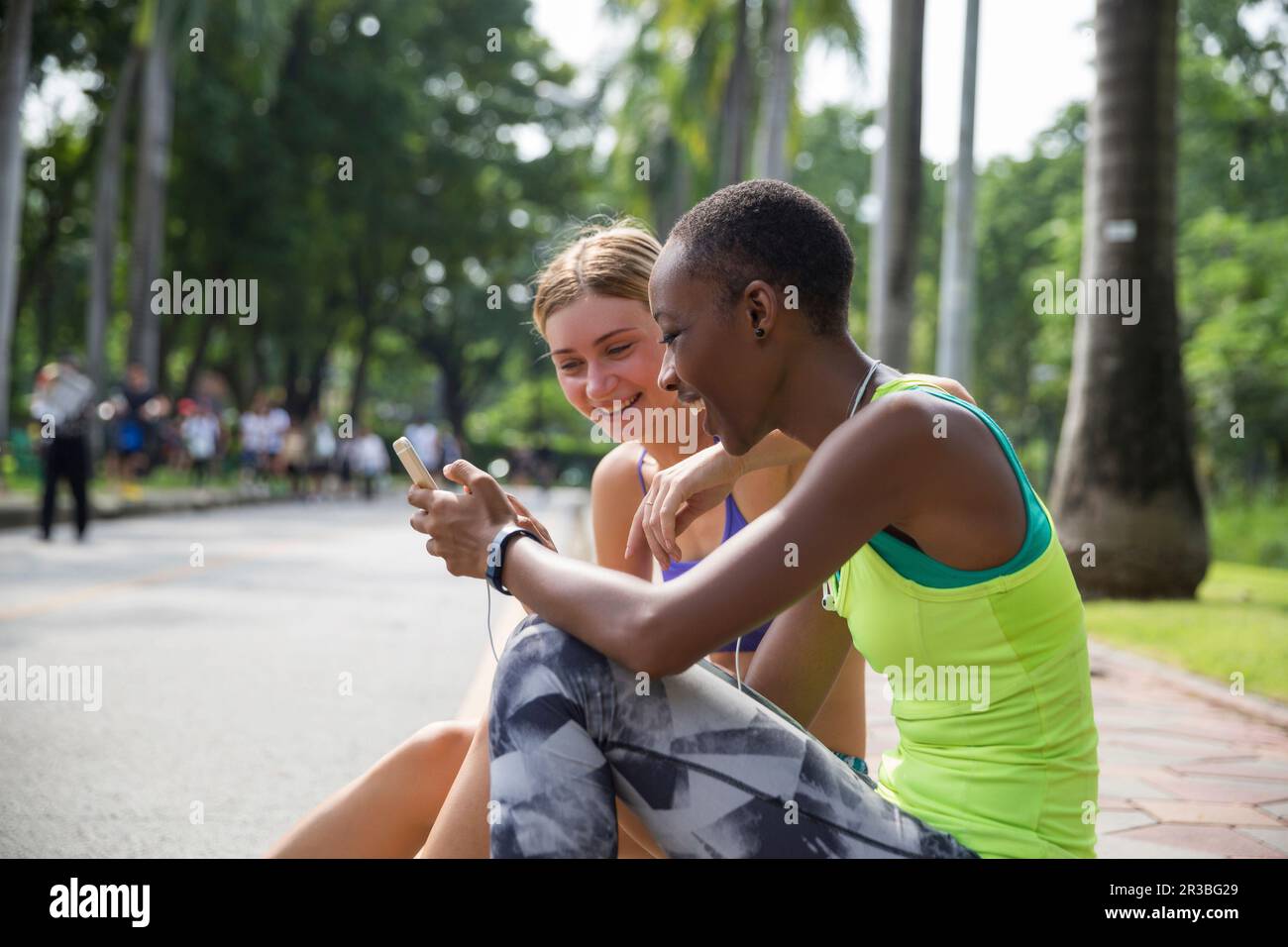 Glückliche Frau mit Smartphone, die mit einem Freund auf dem Bürgersteig im Park sitzt Stockfoto