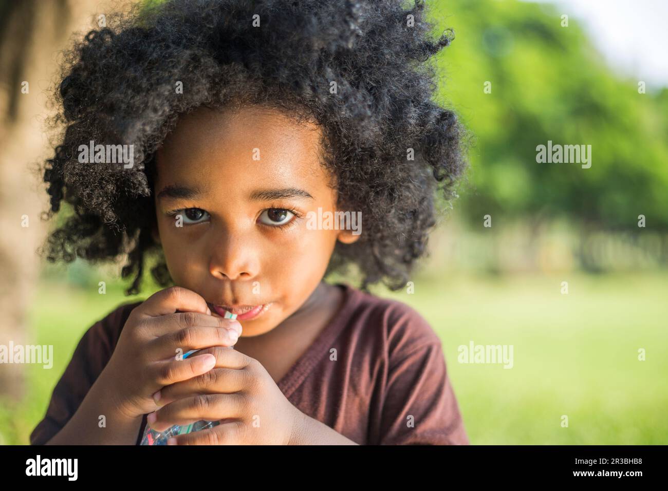Ein Junge mit lockigem Haar, der im Park durch Stroh trinkt Stockfoto