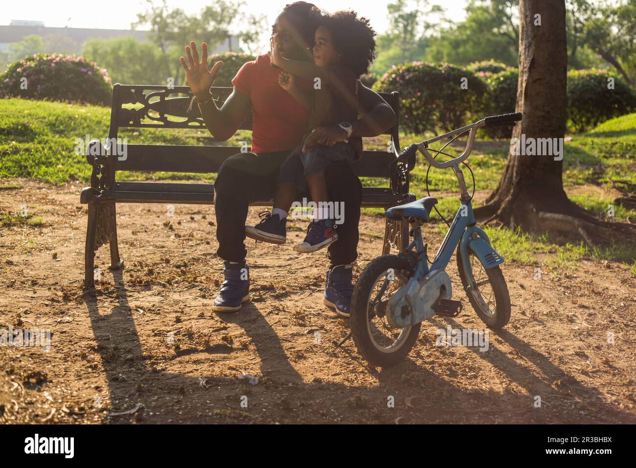 Vater und Sohn winken mit dem Fahrrad, sitzen auf einer Bank im Park Stockfoto