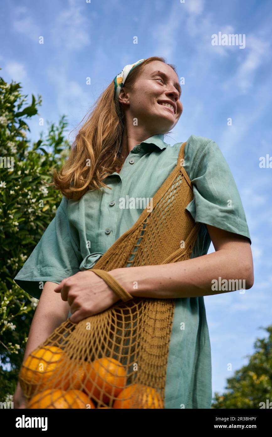 Kontemplative Frau mit Orangen in einem Netzbeutel im Obstgarten Stockfoto