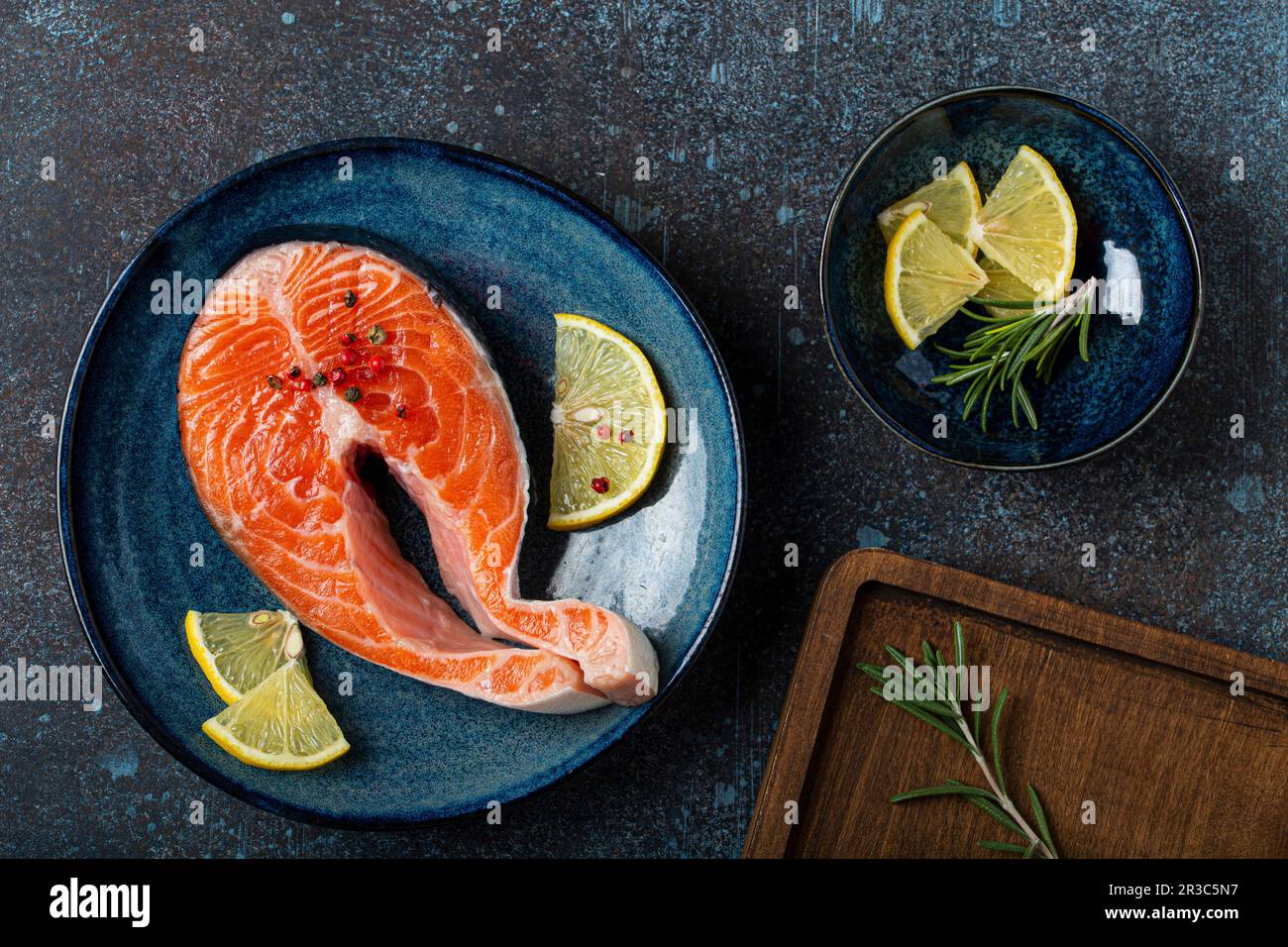 Roher frischer Fisch, Lachssteak, Blick auf Teller, rustikaler dunkler Betonstein-Hintergrund mit Rosmarin, Zitronenspalten und Gewürzen Stockfoto