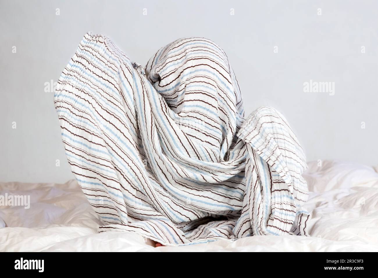 Kleiner weißer Junge, der unter einer gestreiften Decke auf weißer Bettwäsche spielt Stockfoto