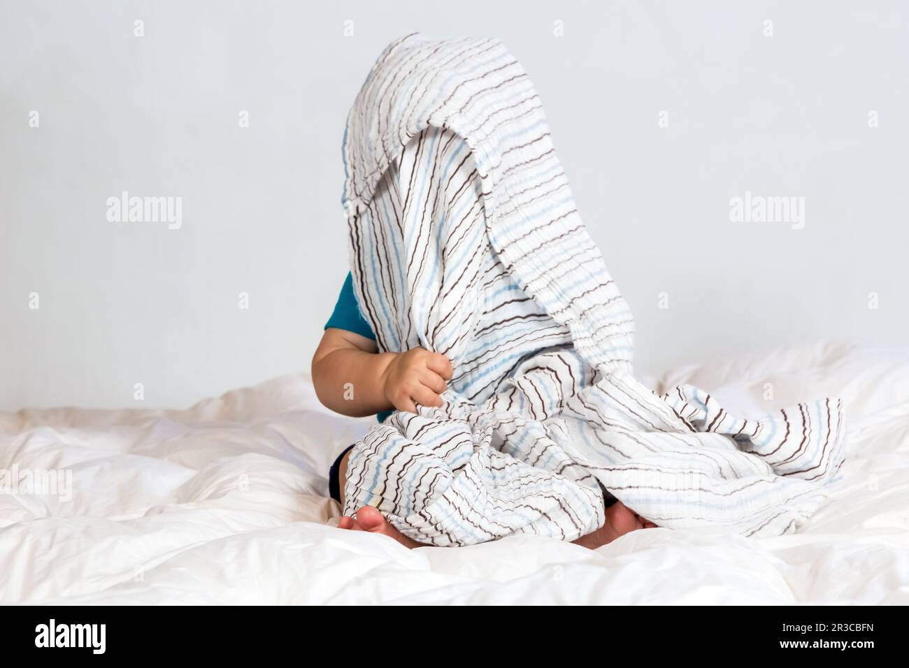 Kleiner weißer Junge, der unter einer gestreiften Decke auf weißer Bettwäsche spielt Stockfoto