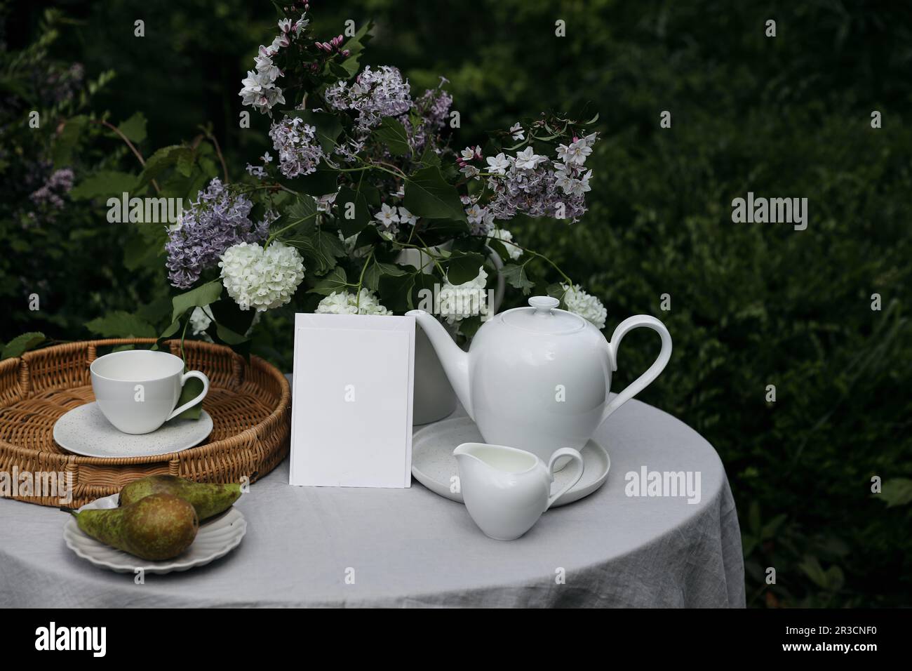 Nahaufnahme einer leeren Grußkarte, ein Geburtstagseinladungs-Modell. Teeparty im grünen Garten. Teekanne, Milchkrug, Birnenfrucht auf dem Tisch mit Leinen Stockfoto