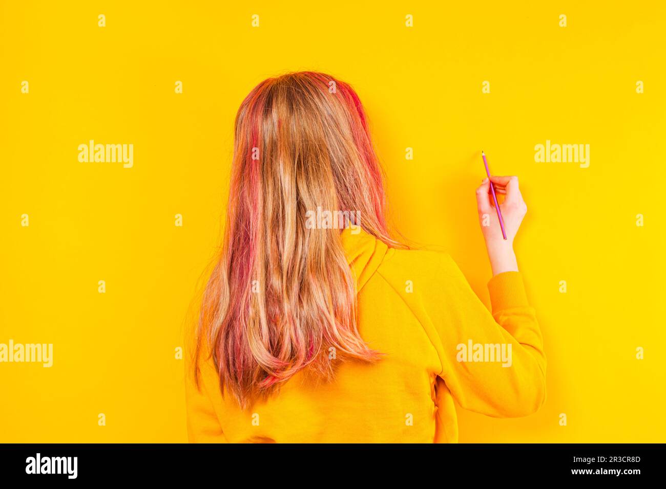 Rückansicht eines jungen Mädchens, das sich auf einen gelben Hintergrund vorbereitet. Das blonde Mädchen in einer gelben Jacke hat einen Bleistift Stockfoto