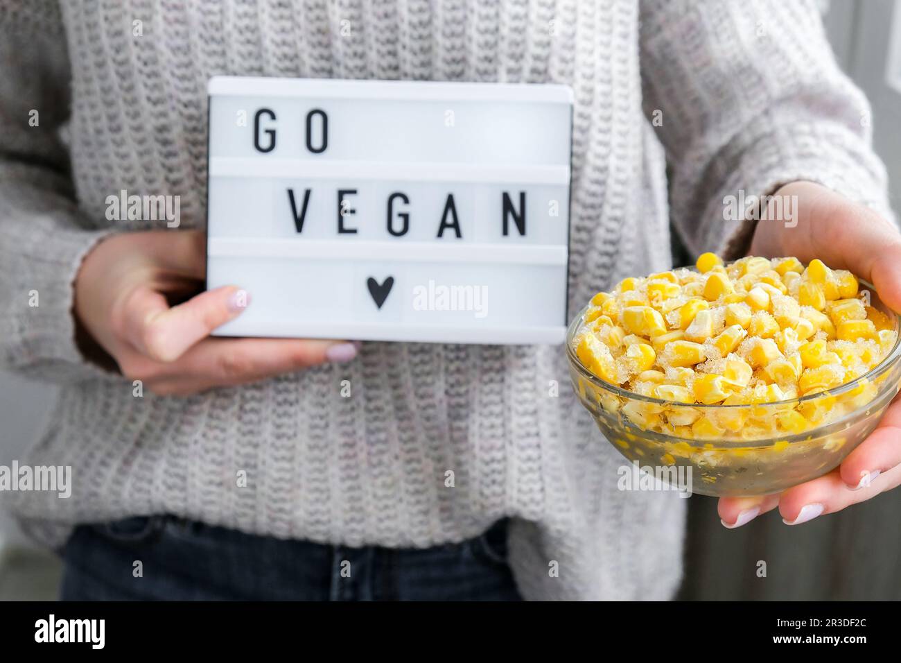 Lightbox mit Text GEHEN VEGAN in weibliche Hände. Veganismus, vegetarisch gesunder Lebensstil. Gefrorene Lebensmittel in der Schüssel. Gelber Mais Stockfoto