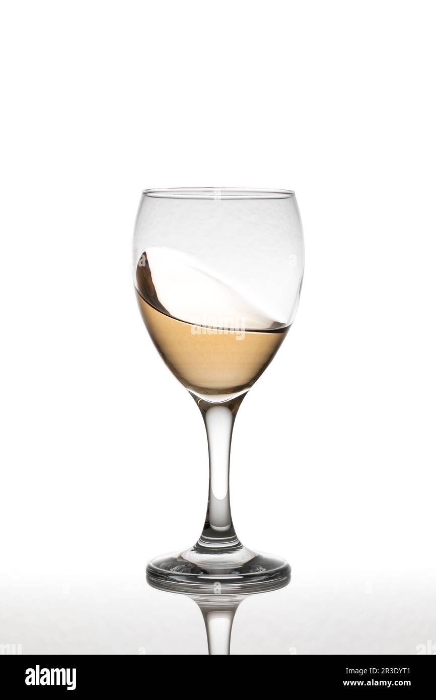 Ein Glas mit Weißwein, das sich über eine reflektierende Oberfläche bewegt und eine sanfte Welle auf weißem Hintergrund zeigt Stockfoto