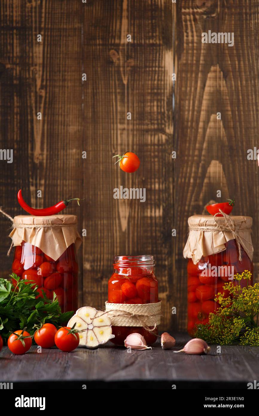 Hausgemachte Tomaten in Dosen. Tomaten fallen in ein Glas auf einem Holzhintergrund mit Platz für Text. Ein offenes Gefäß mit Kirschtomaten, die hinein fliegen. Stockfoto