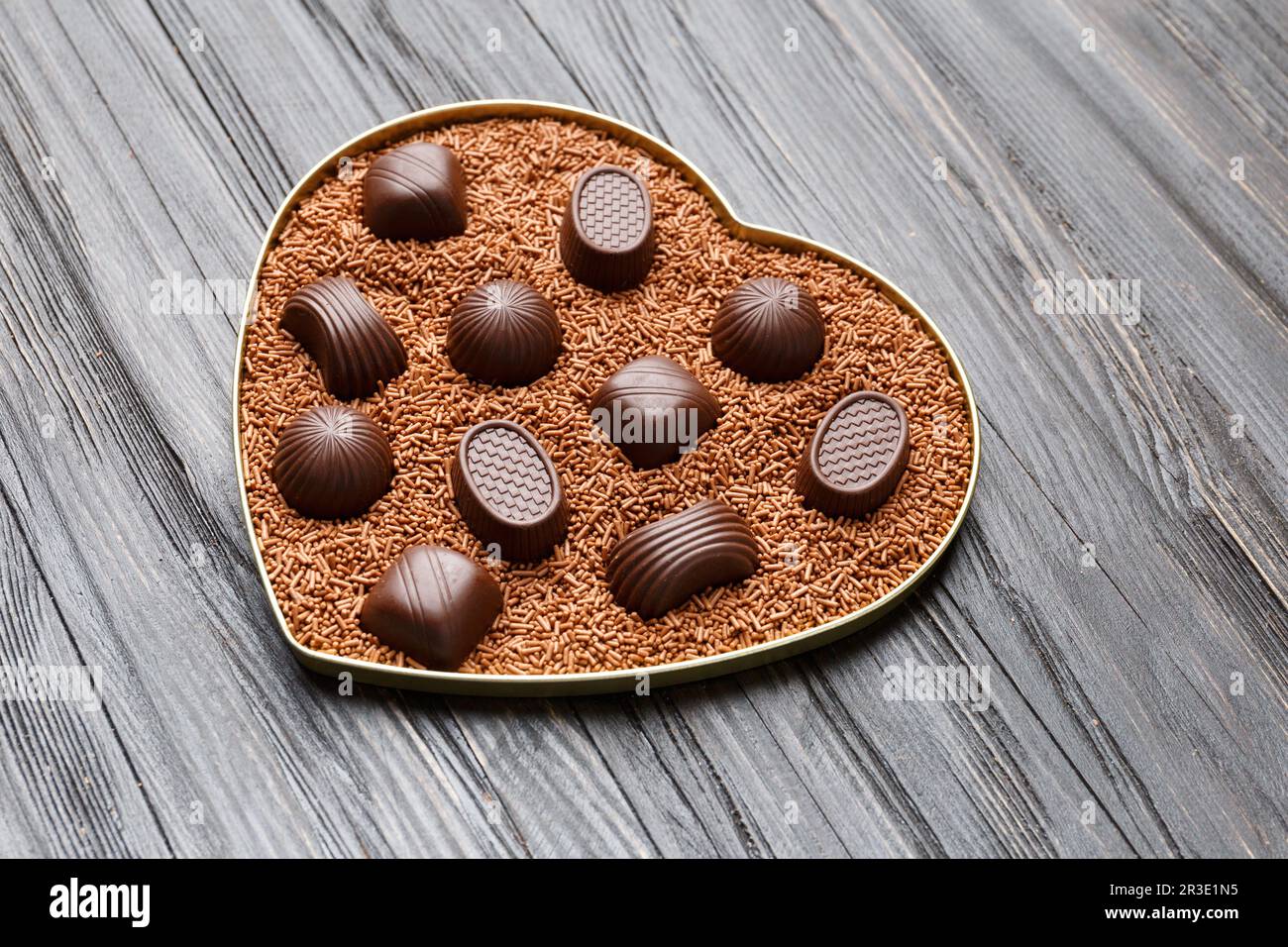 Schokoladenbonbons in Schokoladenstückchen in einer herzförmigen Schachtel auf einem dunklen Holzhintergrund mit Blick auf den Valentinstag. Stockfoto