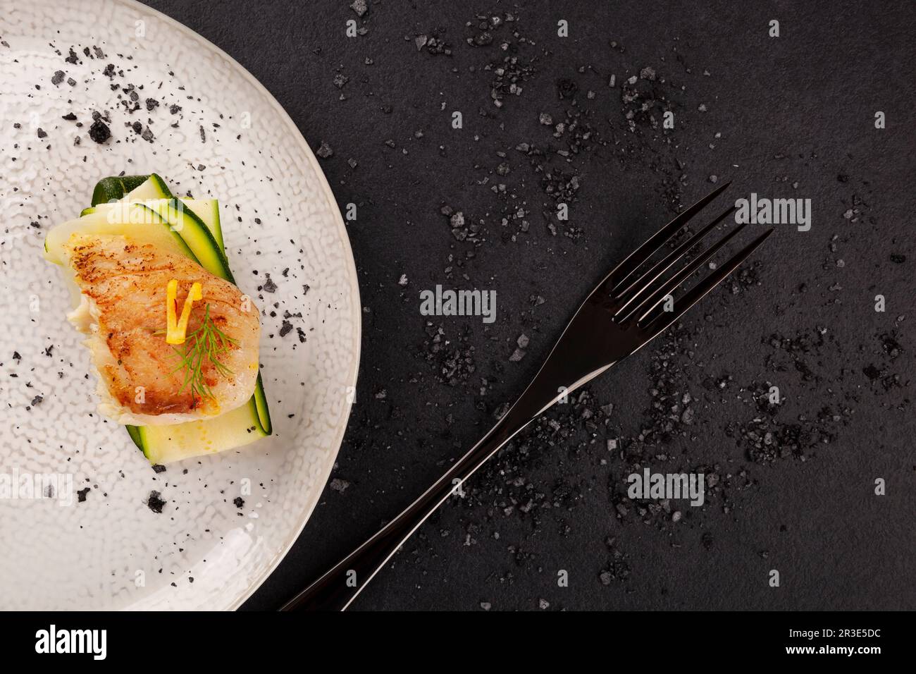 Gegrilltes Fischfilet mit Zucchini auf weißem Teller auf schwarzem Hintergrund. Stockfoto