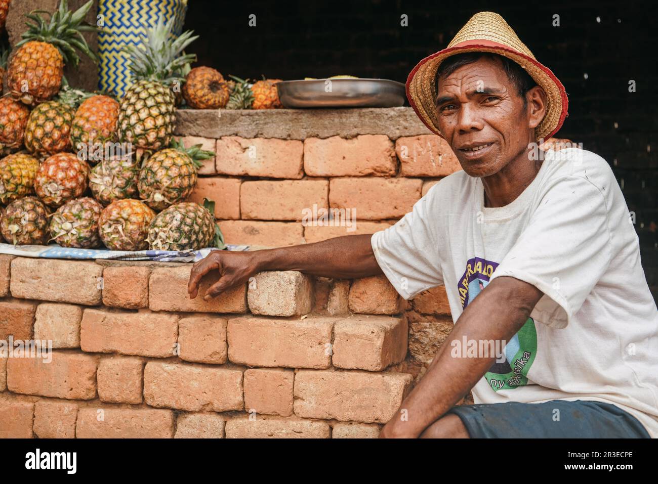 Tsiafahy, Madagaskar - 25. April 2019: Einheimischer madagassischer Mann mit Strohhut und Hemd, der Ananas auf dem Markt verkauft, in die Kamera schaut, Früchte in seiner Nähe Stockfoto