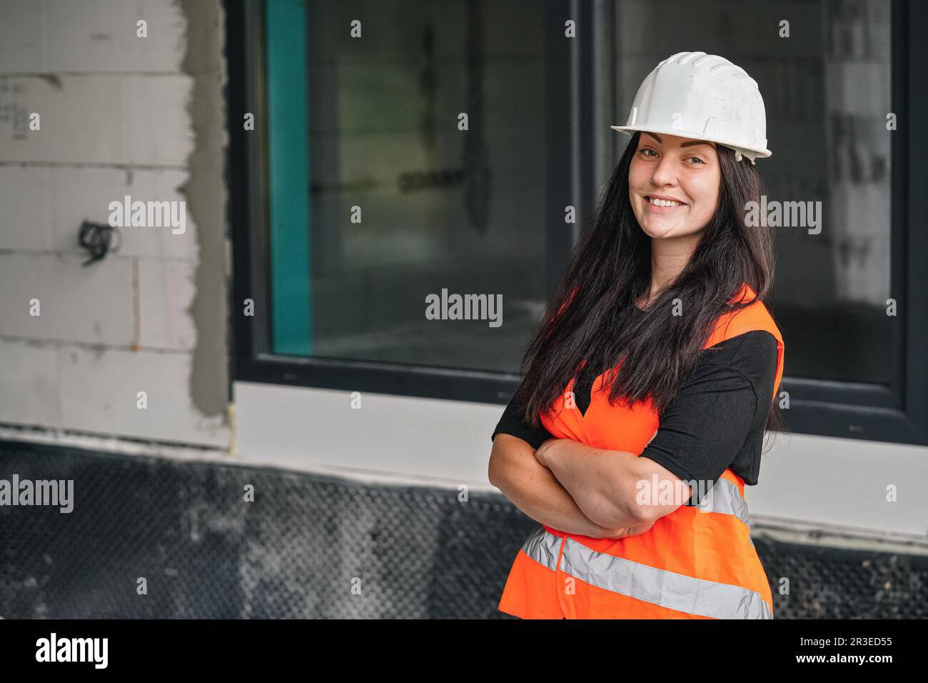 Junge Frau mit weißem Schutzhelm und orangefarbener Warnweste, langes dunkles Haar, Blick in die Kamera, Hände zuversichtlich gekreuzt. Unscharfe Baustelle Stockfoto