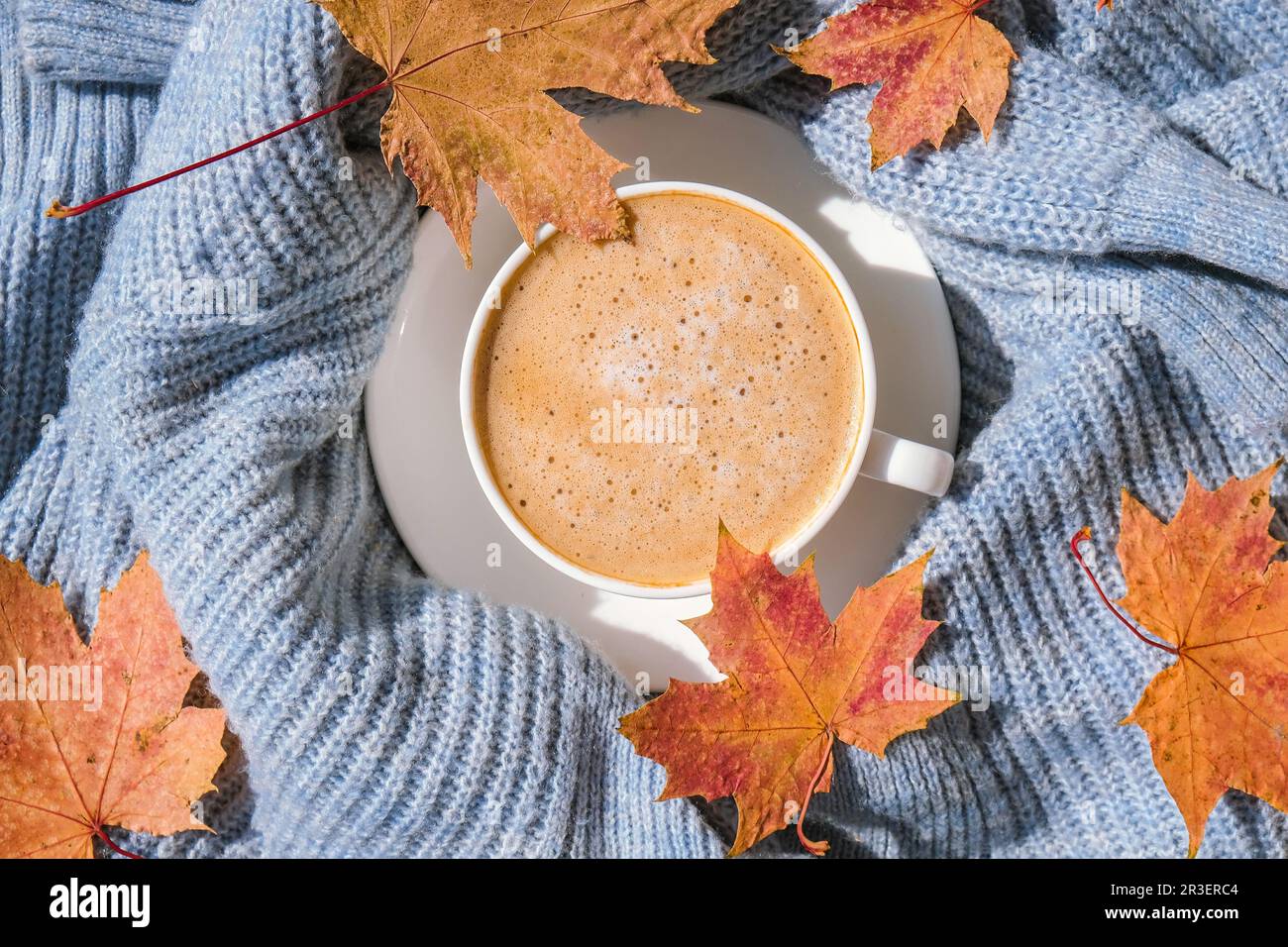 Weiße Tasse Kaffee zum Aufwärmen am Morgen auf blauem Strickpullover mit aphorngelben Blättern auf dem Hintergrund. Gemütliches Hauskonzept. Ästhetik b Stockfoto