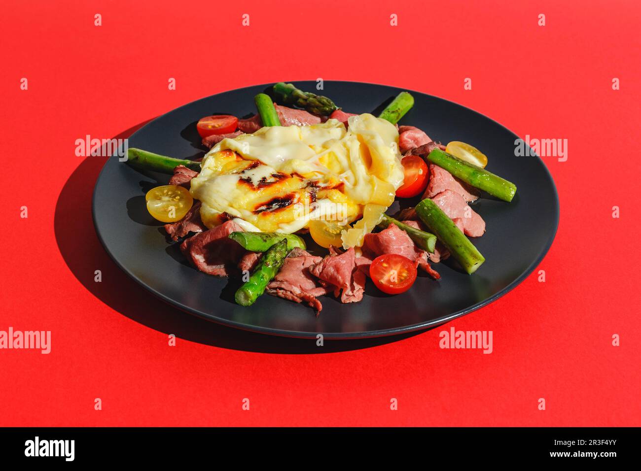 Köstlicher Raclette-Käse, serviert über Rinderbraten mit Tomaten und Spargel, Gericht auf rotem Hintergrund. Hartes Licht, tiefer Schatten Stockfoto