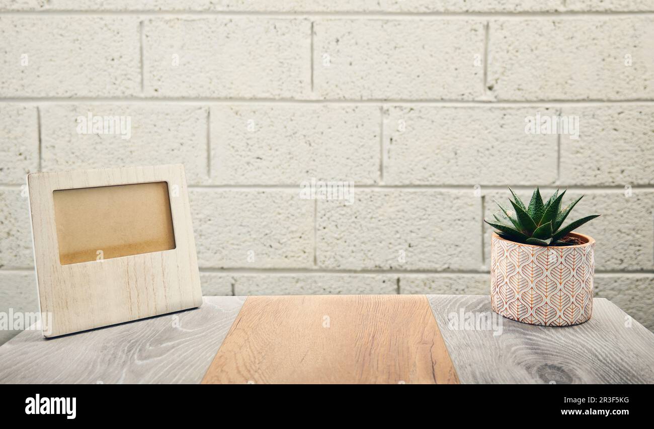 Konzeptaufnahme eines leeren Holzbilderrahmens auf einem Holzschreibtisch mit einer Zimmerpflanze und einer Backsteinwand im Hintergrund Stockfoto