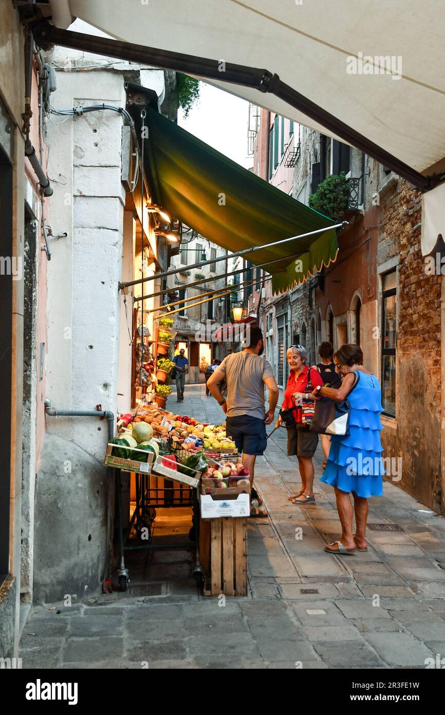 Zwei Damen, die sich mit einem Gemüsehändler vor seinem Obst- und Gemüseladen unterhalten, in einer engen Gasse in der Sestiere von Cannaregio, Venedig, Venetien, Italien Stockfoto