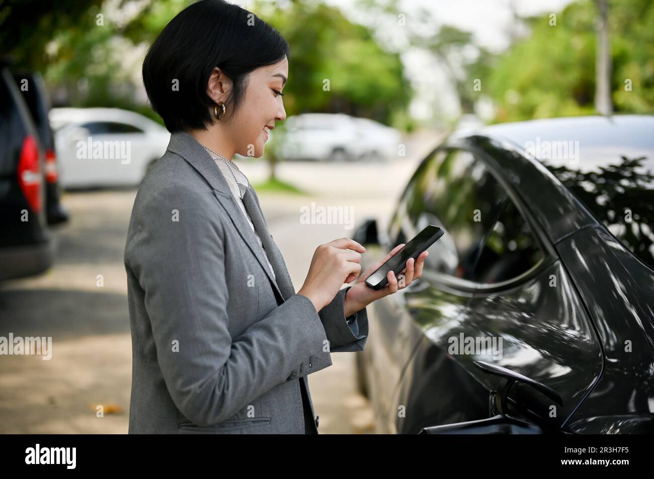 Eine erfolgreiche asiatische Geschäftsfrau liest Nachrichten auf ihrem Smartphone ab, während sie darauf wartet, dass ihr Elektroauto an einer öffentlichen Ladestation vollständig aufgeladen ist. Stockfoto