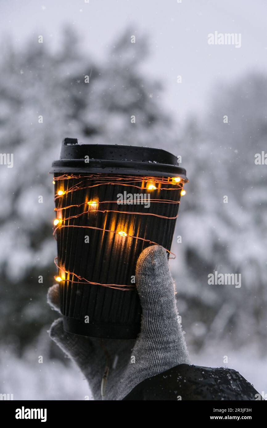 Weibliche Hand in Winterhandschuhen mit schwarzem Öko-Papierbecher und glänzendem Girlande-Licht. Kreativer, trendiger recycelbarer Becher ohne Abfall. Stockfoto