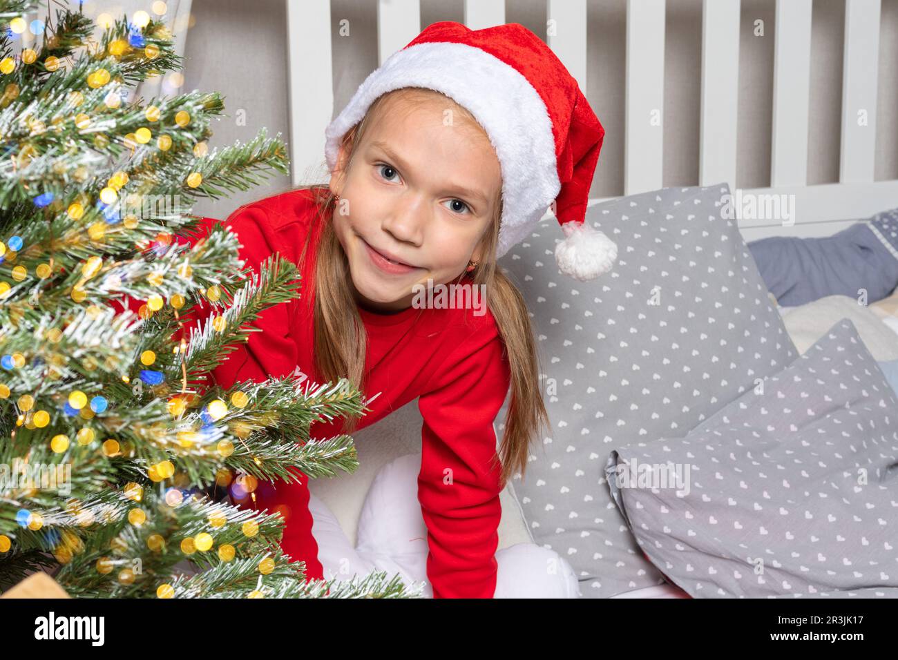 Ein süßes Teenager-Mädchen im roten Pyjama und einem Weihnachtsmann-Hut sieht einen Weihnachtsbaum mit Bokeh-Lichtern auf dem Bett an. Silvester-Morgen. Weihnachtsfamilie Stockfoto