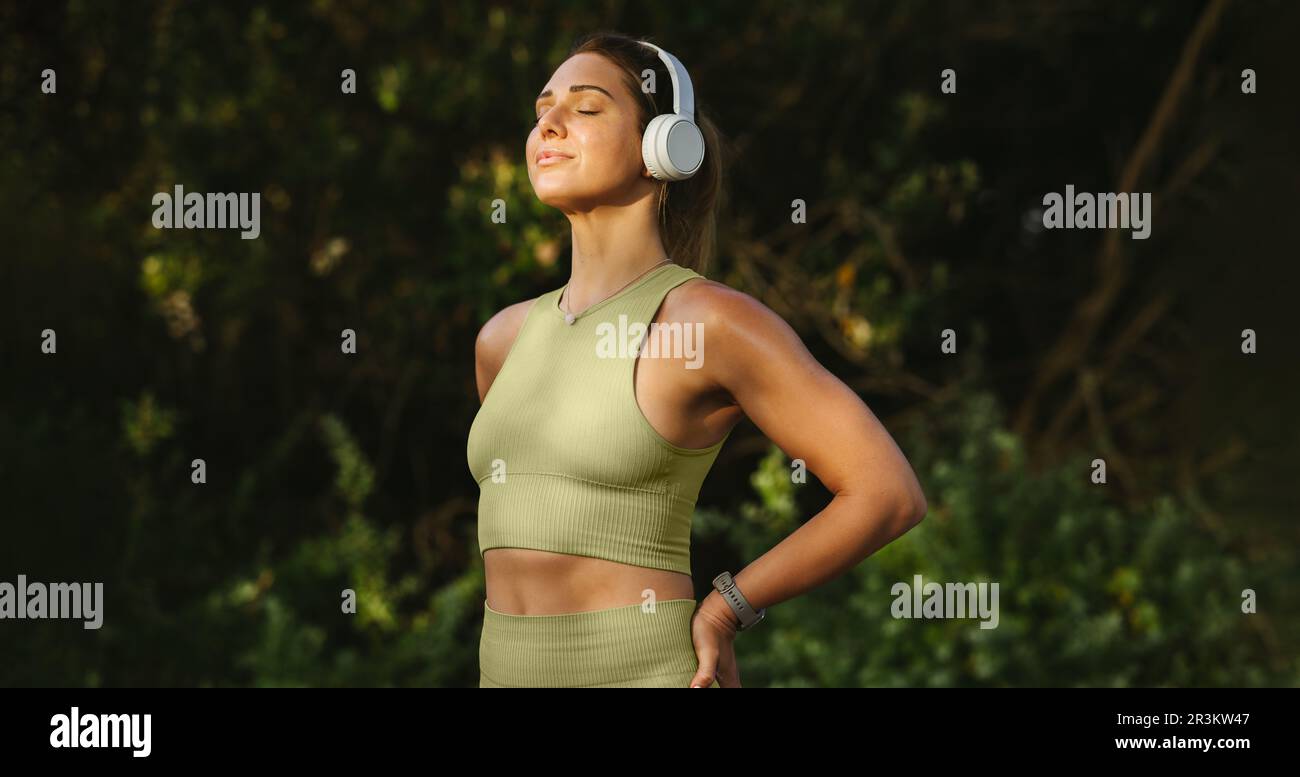 Frau, die Musik hört und draußen eine Atemübung macht. Frau meditiert, während sie vor einem Naturhintergrund steht. Sportlerin wärmt sich auf Stockfoto