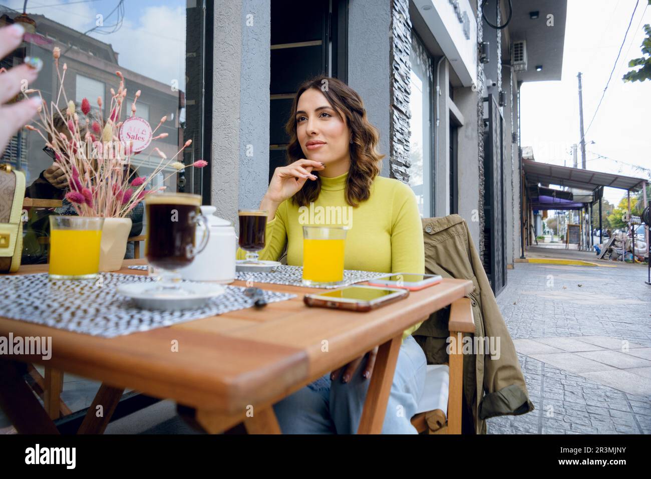 Junge argentinische latina, angezogen in lässiger Kleidung, sitzt vor der Cafeteria und hört ihrem Freund zu, wartet auf das Frühstück Stockfoto