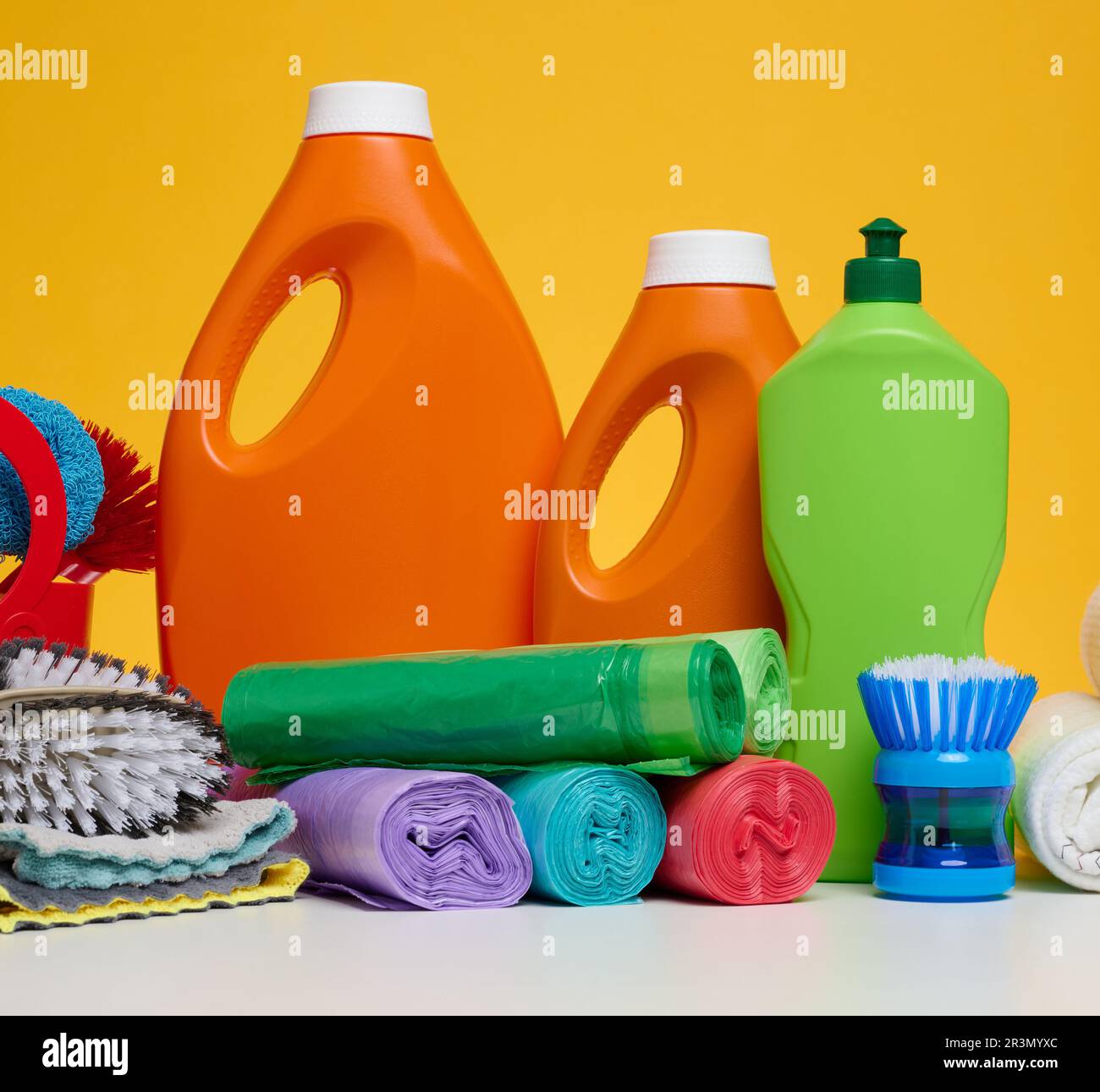 Orangefarbene Kunststoffflaschen mit flüssigen Produkten und Müllsäcken auf einem weißen Tisch, gelber Hintergrund Stockfoto