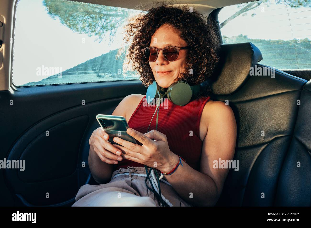Erwachsene ethnische Frau mit lockigem Haar in Sonnenbrille und Kopfhörern, die im Auto auf dem Rücksitz auf ein Mobiltelefon schaut Stockfoto