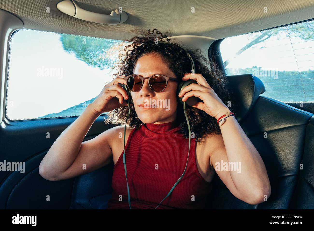 Erwachsene, ethnische Frau mit lockigem Haar in Sonnenbrille und rotem Kopfhörerdesign, während sie im Auto auf dem Rücksitz Musik hört Stockfoto