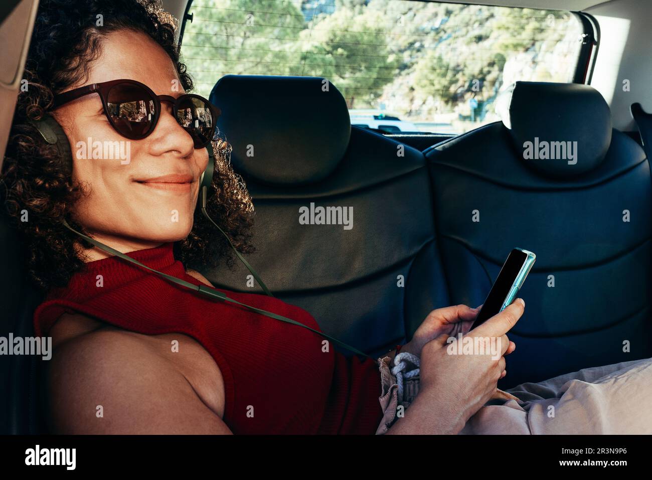Seitenansicht einer erwachsenen ethnischen Frau mit lockigem Haar in einer Sonnenbrille mit Smartphone im Auto, die auf dem Rücksitz sitzt und Musik hört Stockfoto