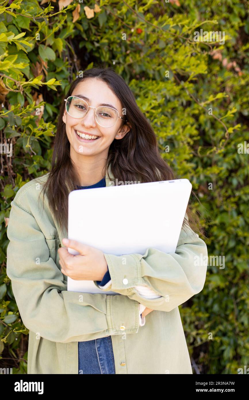 Positive junge Frau in lässiger Kleidung, die am Tag mit einem Laptop gegen den grünen Busch steht und lächelt und in die Kamera schaut Stockfoto