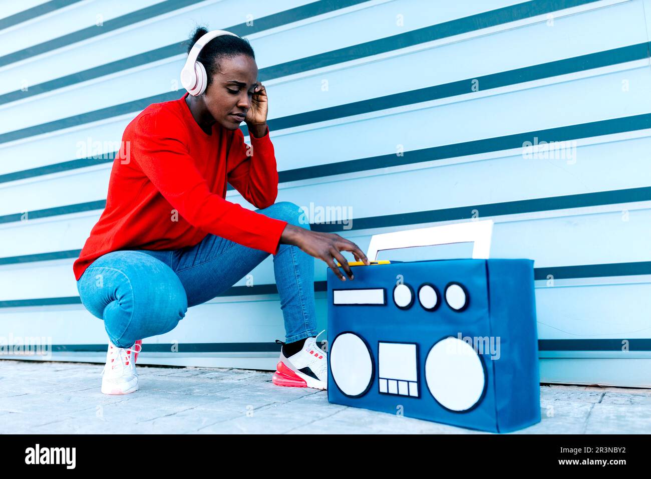 Ein komplettes afroamerikanisches weibliches Modell in Kopfhörern, das eine Playlist mit flippigen Liedern hört, während es neben der Boombox auf dem Bürgersteig sitzt Stockfoto