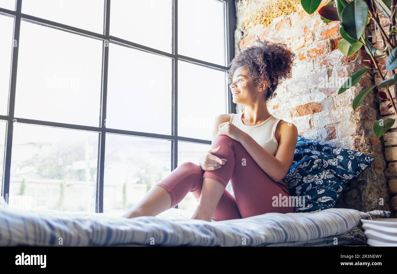 Eine entspannte, afroamerikanische Ethnikerin in Haushaltskleidung, die auf einer Fensterbank sitzt Stockfoto Eine entspannte, afroamerikanische Ethnikerin in Haushaltskleidung, die auf einer Fensterbank sitzt Stockfoto