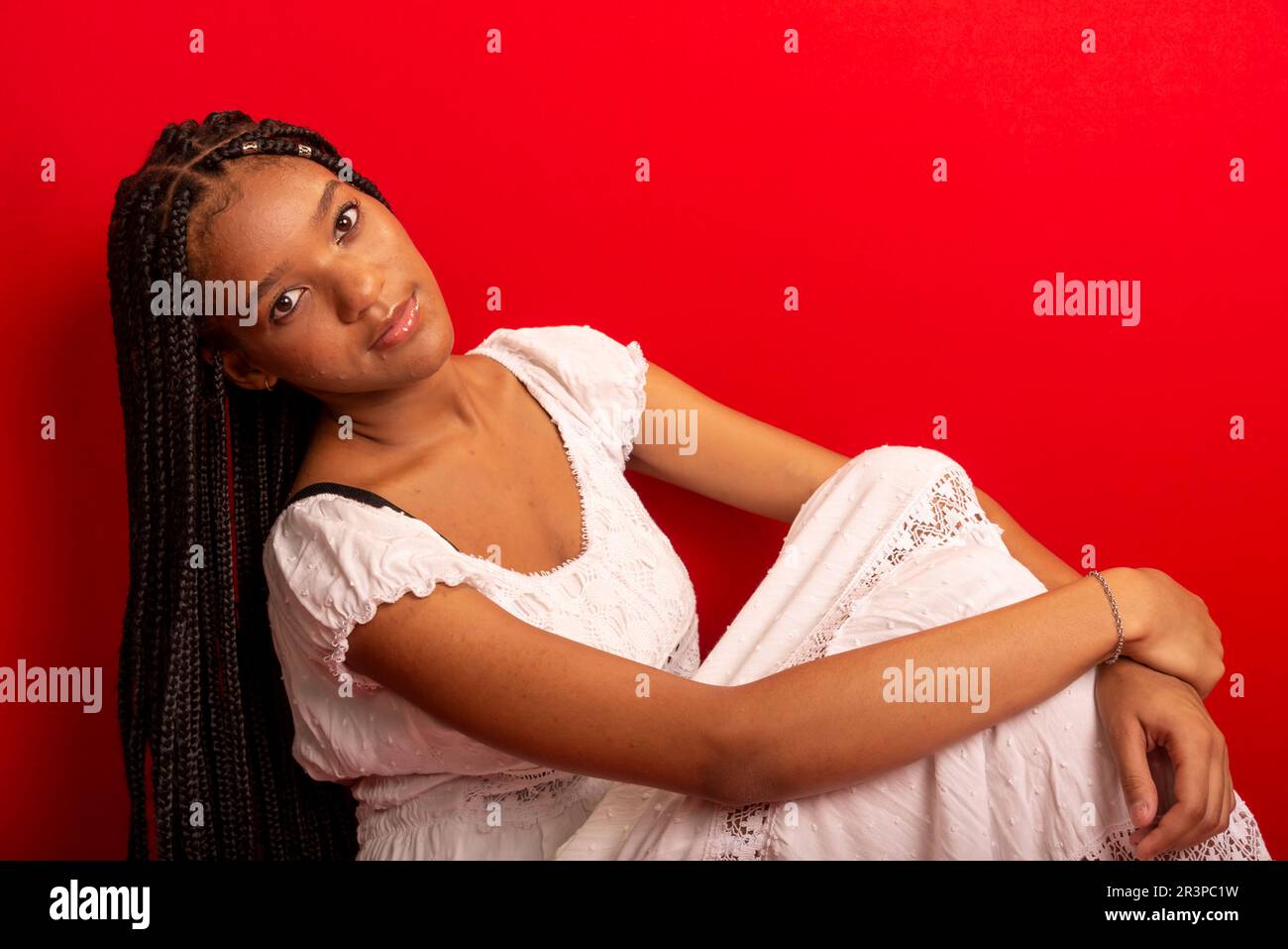 Schöne junge schwarze Frau, die ruhig sitzt und weißes Outfit mit langen Zöpfen trägt. Isoliert auf rotem Hintergrund Stockfoto